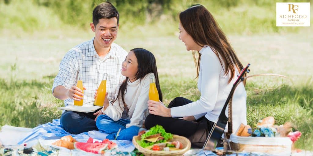  A family sitting at a picnic table, enjoying a meal together. They are eating sandwiches, chips, and fruit.